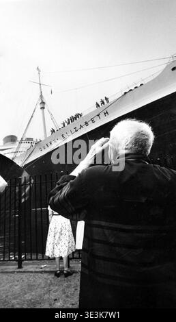 Dagli anni '1950, questa fotografia in bianco e nero di Angelo Rizzuto cattura un uomo più anziano, visto da dietro, guardando verso il maestoso transatlantico RMS Queen Elizabeth. La nave è ormeggiata in quella che è presumibilmente New York City, con il suo nome chiaramente visibile sullo scafo, mentre un assaggio della moda anni '1950 è offerto da una donna in una gonna a pois in primo piano. Foto Stock