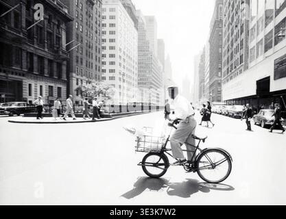 Questa straordinaria fotografia in bianco e nero di Angelo Rizzuto, del 1950, cattura una scena quotidiana di un uomo non identificato in bicicletta con un cestino di consegne attraverso Park Avenue a New York City. La vivace strada è costeggiata da auto e pedoni classici della metà del secolo, che mostrano la moda e l'ambiente urbano dell'epoca. Foto Stock