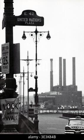 Questa fotografia di strada in bianco e nero degli anni '1950 cattura una scena di strada a New York, di Angelo Rizzuto. Presenta un cartello stradale E. 42 United Nations Plaza, lampioni d'epoca e un cartello "Curb Dog Violation $25 fine", con un taxi sfocato ed edifici industriali sullo sfondo. Foto Stock