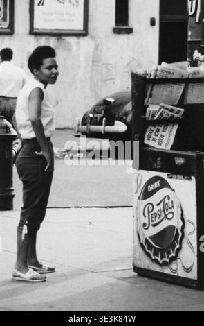 Questa accattivante fotografia di strada in bianco e nero di Angelo Rizzuto, probabilmente degli anni '1950, cattura una giovane donna in piedi accanto a un'edicola di New York City. L'immagine mostra splendidamente la vita di tutti i giorni e la moda informale dell'epoca, con titoli visibili dall'edicola e una pubblicità Pepsi-Cola che si aggiunge alla scena Street vintage. Foto Stock