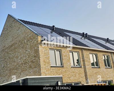 Moderna casa cittadina residenziale in mattoni con pannelli solari installati sul tetto inclinato, caratterizzata da tegole scure e tubi di ventilazione multipli neri. Foto Stock