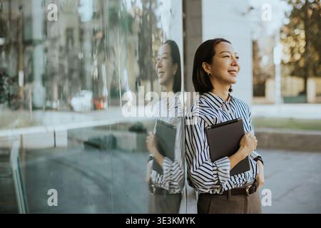 Una donna di origine coreana sorge fuori da un edificio moderno. Tiene una cartella e sorride mentre guarda il suo riflesso nel vetro. E' giorno Foto Stock
