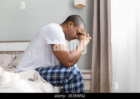 Giovane uomo nero seduto sul letto con le mani schiacciate e testa giù Foto Stock