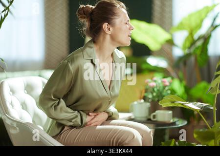 Una donna con una camicia di seta verde chiaro stringe l'addome con entrambe le mani, mostrando segni di intenso dolore allo stomaco o disagio mentre è seduta sulla sua liv Foto Stock