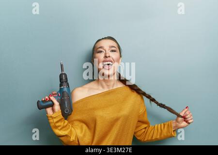 Concetto di fiducia. Donna attraente che tiene trapano e treccia. Espressione forte e sorridente. Stile casual e alla moda. Faccia evidenziata in verticale e AT Foto Stock