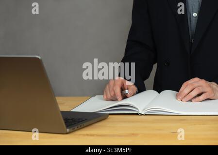 Un uomo è in piedi dietro un tavolo e guarda il suo laptop. Nella mano destra tiene una penna. Sul tavolo giace un quaderno aperto. Foto Stock