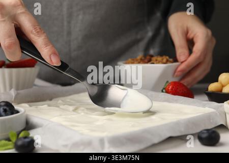 Preparare la corteccia dello yogurt. Donna che spalma lo yogurt con un cucchiaio sul piatto a tavola bianca, primo piano Foto Stock