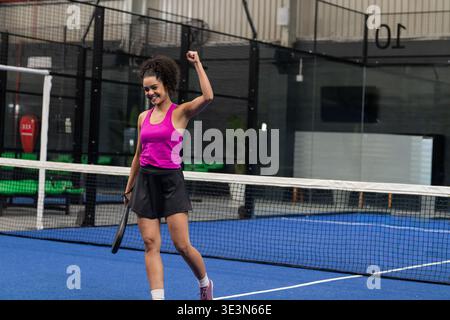 Donna che tiene la racchetta e solleva il pugno, festeggia sul campo blu vicino alla rete, indossa una canotta rosa. Racquet, femminile, atletica, trionfo, struttura, scherma, g Foto Stock
