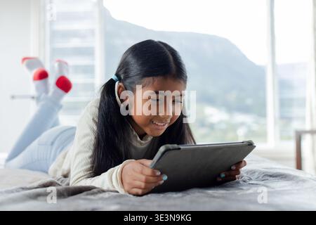 Ragazza in età scolare che usa un tablet nero mentre si sdraia sul letto con una coperta grigia in camera da letto vicino alla finestra Foto Stock