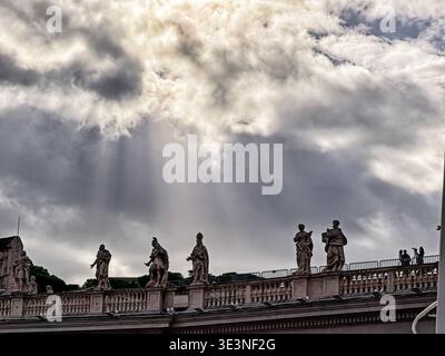 I raggi di Dio emanano da una parte del sole che sgorga attraverso le nuvole sulle statue del colonnato che circonda Piazza San Pietro a Roma. Foto Stock