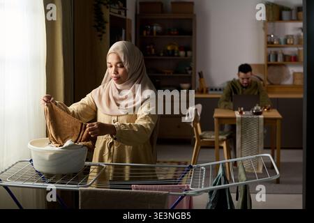 Giovane donna musulmana adulta che piega il bucato vicino alla finestra, uomo mediorientale che lavora su un laptop a un tavolo in background, scena domestica che mostra la vita quotidiana e il multitasking a casa Foto Stock