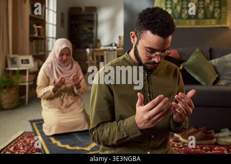 Giovane uomo mediorientale seduto sul pavimento, pregando con le mani alzate di fronte a una giovane donna mediorientale adulta inginocchiata e pregando nel soggiorno decorato con elementi islamici Foto Stock
