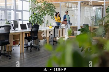 Diverse collaboratrici che camminano accanto alle scrivanie in legno in un ufficio aperto tenendo smartphone e tazza di caffè Foto Stock