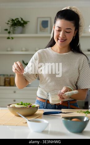 Donna asiatica in piedi in cucina per preparare insalate, spolverare semi dal vaso di vetro Foto Stock