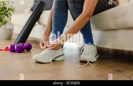 Donna afroamericana che lega i lacci bianchi sul divano notando i manubri che preparano l'allenamento Foto Stock