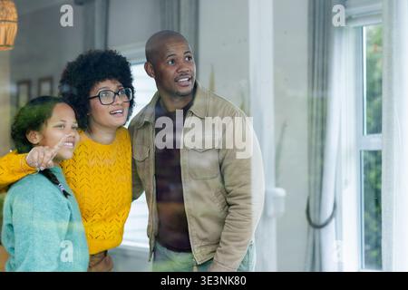 Famiglie diverse che si affacciano attraverso una grande finestra nel soggiorno, incorniciata da tende e lampada a sospensione Foto Stock