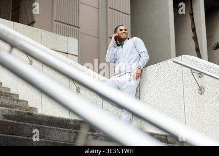 Uomo afroamericano in felpa con cappuccio appoggiato da una scala alla porta che sorride lo smartphone Foto Stock