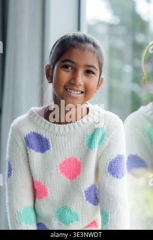 Ragazza asiatica sorridente, seduta a casa accanto alla finestra in maglione bianco con pois, fascia gialla Foto Stock