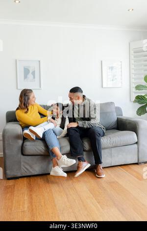 Famiglia di tre persone appoggiate insieme su un divano grigio nel soggiorno di casa con persiane per piantagioni Foto Stock