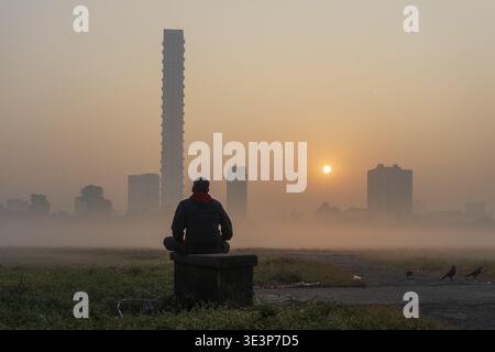 La vista di una figura solitaria si trova in serena contemplazione mentre il sole sorge su un paesaggio nebbioso con edifici torreggianti, Kolkata, Bengala Occidentale, India. Foto Stock