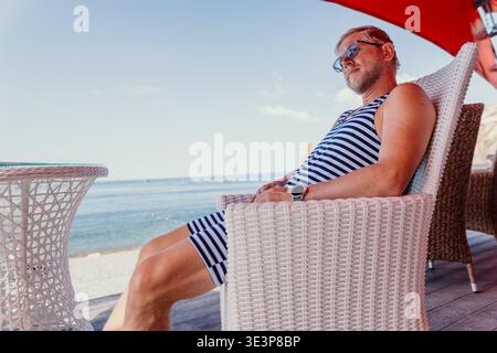 Uomo relax sulla spiaggia l'anziano uomo che indossa una tuta a righe e occhiali da sole siede su una sedia in vimini bianca con vista sull'oceano in un giorno d'estate Foto Stock