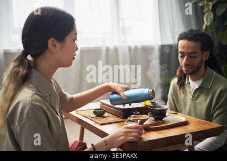 Giovane donna asiatica che versa tè caldo da thermos in tazza mentre siede a tavola con un giovane adulto nero, partecipando alla tradizionale cerimonia del tè in un luminoso ambiente interno Foto Stock