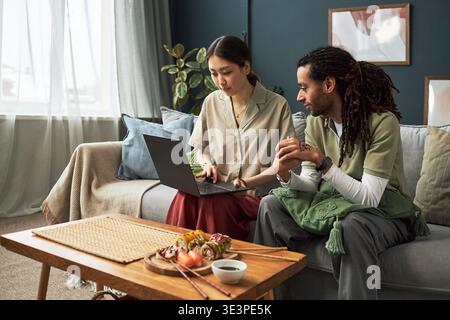 Giovane donna asiatica seduto sul divano usando un computer portatile, mentre il giovane adulto caucasico con i dreadlock seduto accanto a lei tenendosi per mano insieme, piatto di sushi sul tavolo in primo piano Foto Stock