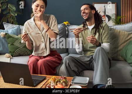 Giovane donna asiatica adulta e giovane uomo nero adulto seduto sul divano ridendo e mangiando con le bacchette mentre guardano il portatile, condividendo momenti di gioia e gustando insieme la cucina asiatica Foto Stock