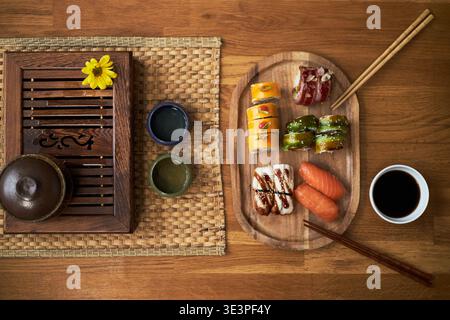 Cucina asiatica tradizionale con pezzi assortiti di sushi, disposti su un piatto di legno con salsa di soia e bacchette accanto al set da tè su tappeto intrecciato che mette in risalto elementi culturali da pranzo Foto Stock