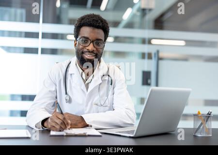Medico di sesso maschile che lavora presso una scrivania affollata della clinica, sorridendo e scrivendo appunti mentre sono visibili un computer portatile e uno stetoscopio, che rappresentano una cura dedicata Foto Stock