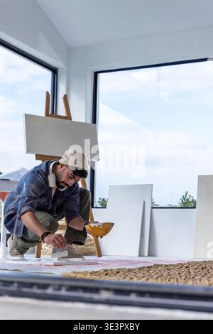 Uomo di 20 anni che indossa un berretto inginocchiato in un luminoso angolo studio, dipinge la tela del pavimento con le cuffie Foto Stock