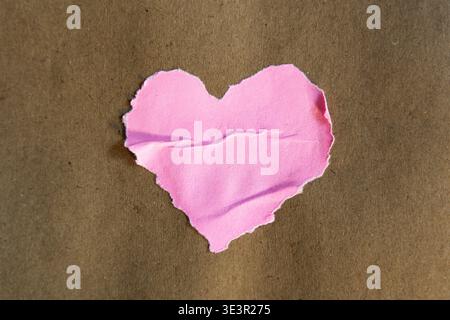 Forma a cuore rosa fatta a mano con bordi strappati e irregolari incollati al centro di uno sfondo rustico di carta marrone. Lo shot cattura il bordo fibroso tattile Foto Stock