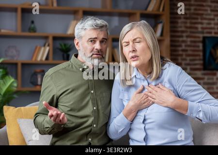 Donna anziana che soffre di grave dolore toracico e difficoltà respiratorie, che stringe il petto, mentre il marito preoccupato guarda avanti con confusione e truffa Foto Stock