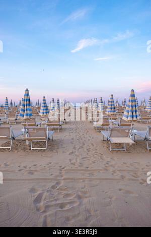 La vista della tranquilla spiaggia si dispiega con file di lettini e ombrelloni a strisce blu che si estendono verso l'orizzonte sotto un cielo pastello, C. Foto Stock