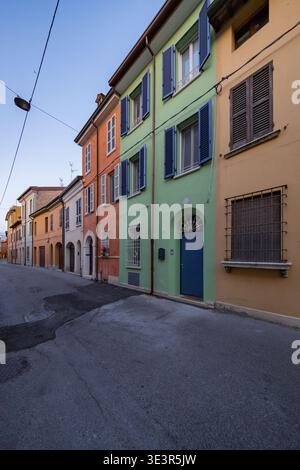 La vista delle case vibranti in tonalità arancione, verde e giallo si staglia fianco a fianco lungo una tranquilla e tortuosa strada asfaltata sotto un cielo limpido, Ravenna, Italia. Foto Stock