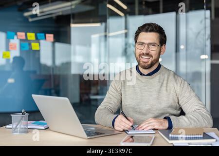 Uomo sorridente con occhiali che lavora a una scrivania, guarda lo spettatore mentre prende appunti in un notebook con un notebook nelle vicinanze in un ufficio moderno, che rappresenta la produttività e il successo aziendale Foto Stock