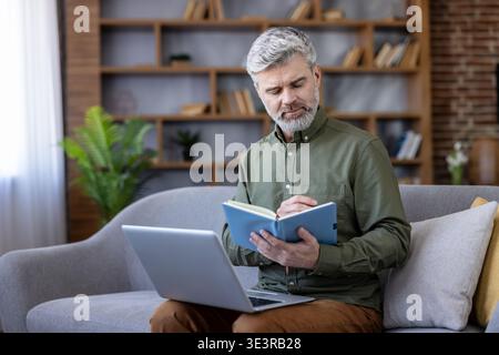 Uomo barbuto dai capelli grigi che lavora da casa su un laptop e un notebook, concentrato e pianifica le attività mentre è seduto comodamente su un divano in un moderno spazio di lavoro in salotto Foto Stock