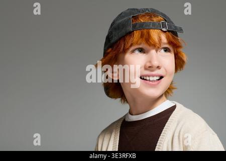 Redhead boy vestito con abiti casual sorride brillantemente mentre posa in un moderno studio. Foto Stock