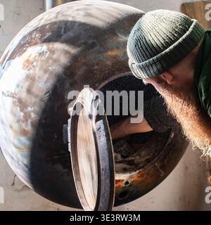 Artigiano barbuti che accende una stufa in metallo fatta a mano all'interno di un laboratorio di lavorazione del legno. Area di lavoro industriale con apparecchiature di riscaldamento utilizzate in un ambiente rustico Foto Stock