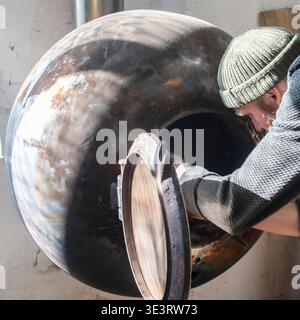 Artigiano barbuti che accende una stufa in metallo fatta a mano all'interno di un laboratorio di lavorazione del legno. Area di lavoro industriale con apparecchiature di riscaldamento utilizzate in un ambiente rustico Foto Stock
