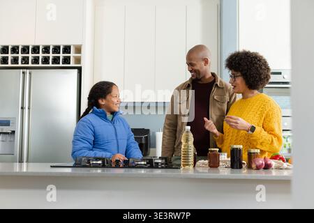 Famiglia diversificata che si trova a Kitchen Island per preparare i pasti utilizzando una stufa a gas integrata e vasetti di spezie Foto Stock