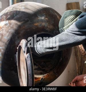 Artigiano barbuti che accende una stufa in metallo fatta a mano all'interno di un laboratorio di lavorazione del legno. Area di lavoro industriale con apparecchiature di riscaldamento utilizzate in un ambiente rustico Foto Stock