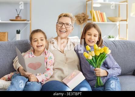 Donna matura sorridente seduta sul divano, abbracciando i bambini congratulandosi con lei con tulipani e carta fatta a mano. Foto Stock