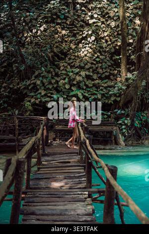 Donna su un ponte di legno sul fiume turchese della giungla circondato da una lussureggiante foresta tropicale Foto Stock
