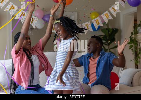 Famiglia afroamericana che fa il tifo sul divano a casa, bambino che salta in abito blu a righe con coriandoli Foto Stock