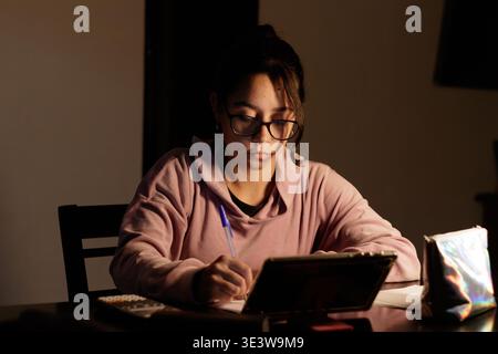 La luce soffusa si illumina sopra il suo volto focalizzato. Scrive con penne e carte, occhi luminosi e intenti. Gli occhiali incorniciano il suo sguardo concentrato. Toni caldi alti Foto Stock