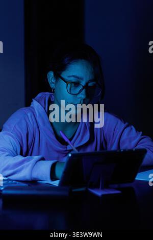 La giovane donna studia intensamente sotto una luce blu fredda. Indossa degli occhiali, concentrata sul suo tablet. La camera oscura enfatizza la sua concentrazione. Istruzione, studio Foto Stock