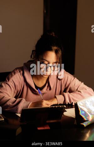 Una giovane donna studia con la messa a fuoco sotto una calda luce da scrivania. Indossa occhiali e una felpa con cappuccio rosa tenue. La sua mano scrive accanto a una tavoletta su una superficie scura. Met Foto Stock
