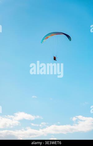Un parapendio si innalza dolcemente in alto attraverso un'ampia distesa di cielo azzurro limpido con nuvole sparse, catturando l'essenza della libertà e dell'avventura Foto Stock