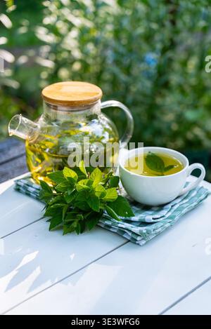 Una rinfrescante scena di tè alla menta fatto in casa, preparato in una teiera di vetro, accompagnato da una tazza, su un tavolo bianco all'aperto. Foglie di menta fresche accentuano la n Foto Stock